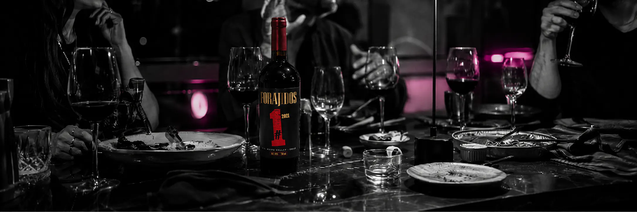 Mesa nocturna compartida con copas, platos y una botella Forajidos